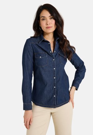 Camisa de mezclilla azul oscura con mangas largas, dos bolsillos frontales, cierre de botones y detalles de costura en contraste. Textura suave y diseño ajustado.