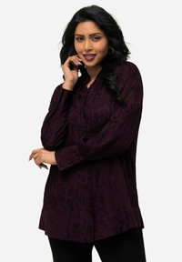 Dark purple blouse with a textured floral pattern, button closure, and ruffle detailing along the neckline and cuffs. Long sleeves.