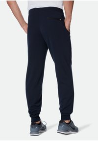 Navy Jogginghosen aus weichem Stoff mit schmal zulaufenden Beinen, seitlichen Taschen und einer Reißverschlusstasche auf der Rückseite. Kombiniert mit grauen Sportschuhen.