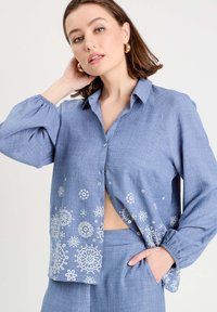Chemise boutonnee bleu clair en tissu texturé, ornée de motifs floraux blancs le long de l'ourlet et présentant une coupe décontractée avec des manches légèrement ballons.