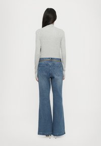 Femme aux longs cheveux foncés, debout de dos, portant un haut moulant gris clair à manches longues et un jean bleu évasé taille haute avec une ceinture chaîne.