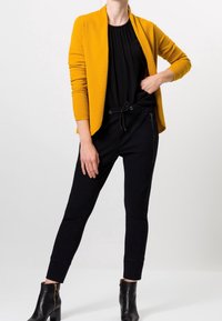 Cárdigan texturizado amarillo sobre una blusa negra, combinado con pantalones negros ajustados que tienen cremalleras laterales y botas negras hasta el tobillo con un acento dorado.