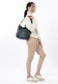 Mustasta nahasta valmistettu pyöreä olkalaukku, jossa hopeanväriset metalliosat ja kohopainettu logo. Laukkua kantaa henkilö, jolla on päällään kerma valkoinen neulepaita ja beiget housut.