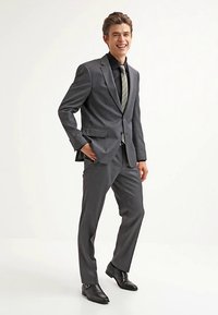 Grijs krijtstreeppak met inkeping revers, gecombineerd met een zwart overhemd en een groen gestreepte stropdas. Smalle broek en zwarte leren schoenen.