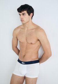 White cotton boxer briefs with a navy waistband featuring a logo. The material is smooth, fitted, and stretches slightly.