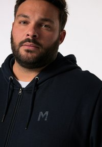 Felpa con zip di un blu navy scuro, con un logo "M" testurizzato sul petto. Dotata di stringhe e di una texture del tessuto morbida e liscia.