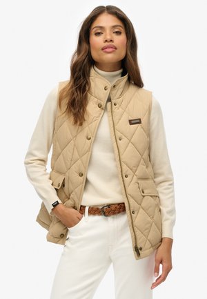 Beige quilted vest with snap buttons, high collar, and side pockets, worn over a light sweater, paired with white pants and a braided belt.