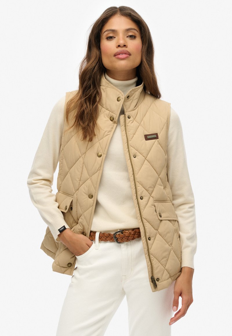 Gilet matelassé beige avec boutons-pression, col haut et poches latérales, porté sur un pull léger, associé à un pantalon blanc et une ceinture tressée.