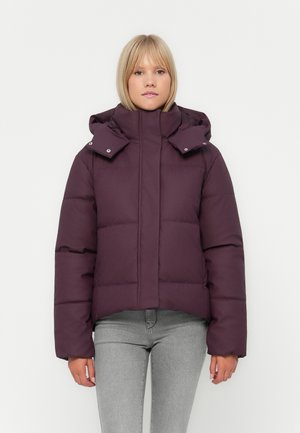 Femme portant une veste d’hiver matelassée violet foncé avec un col montant et une capuche, associée à un jean gris clair, debout sur un fond blanc.