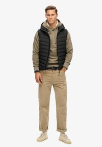 Czarna pikowana kamizelka na beżowym hoodie, khaki spodnie i jasne sneakersy. Stylizacja ma swobodny, warstwowy wygląd.