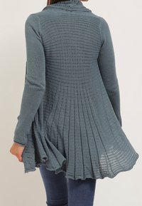 Stickad cardigan i en mjuk blå-grön nyans, med huva, strukturerad mönster och utsvängd design med plisserad baksida och volangkant.