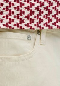 Gros plan sur la poche d'un pantalon en denim beige avec des rivets métalliques sous un bord de tissu tissé blanc et rouge.