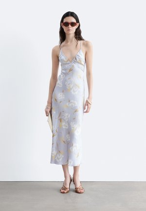 Femme portant une robe nuisette fleurie violet clair, des talons marron à brides, des lunettes de soleil rouges, tenant une pochette, debout contre un mur blanc.