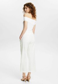 Femme aux cheveux bouclés portant un haut blanc à épaules dénudées, un pantalon large blanc et des sandales à talons beiges, se tenant de profil avec les mains dans les poches.