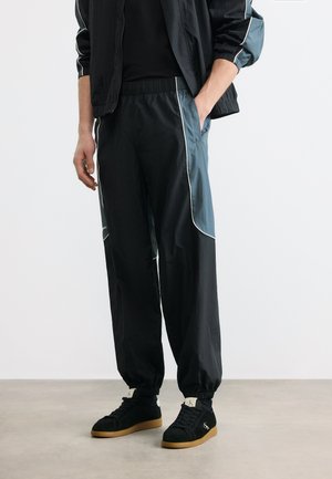 Mees, kes kannab musta ja halli dressipükse valgete torudega, musti tossusid kummeeritud tallaga ning musta jopet, seistes hallil põrandal.