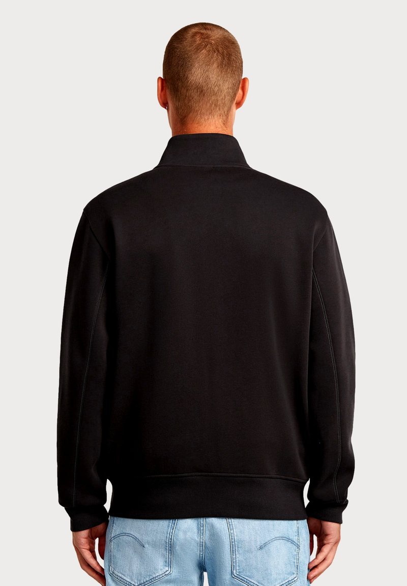 Felpa nera con collo alto, polsini e orlo a coste. Tessuto liscio con cuciture laterali visibili, abbinata a jeans azzurri.