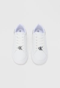 Ζευγάρι λευκά αθλητικά παπούτσια χαμηλού προφίλ με λευκά κορδόνια και μεταλλικά κρεμαστά διακοσμητικά με το λογότυπο "cK" στα κορδόνια κάθε παπουτσιού.