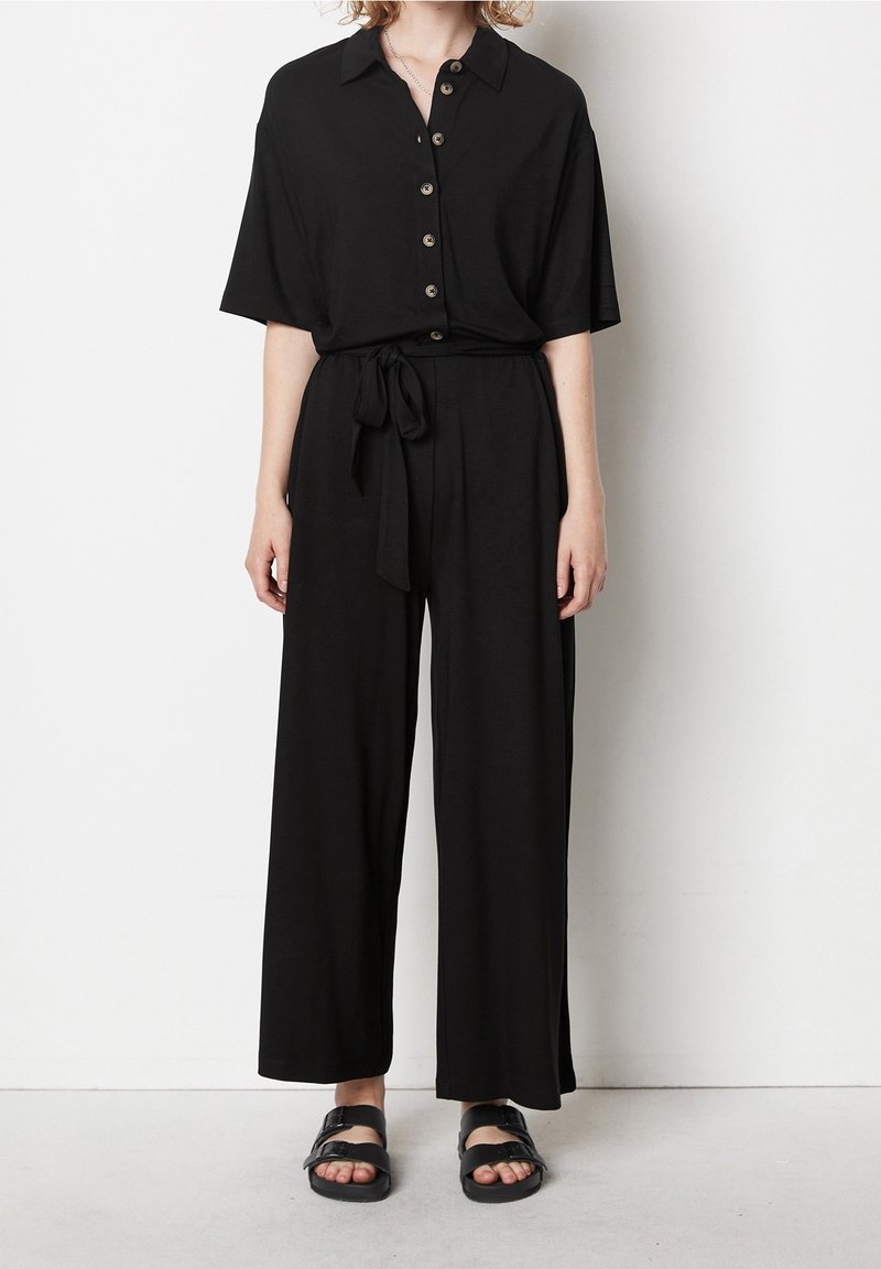 Femme portant un haut noir à manches courtes à boutons avec col et un pantalon large assorti noué à la taille, ainsi que des sandales noires.