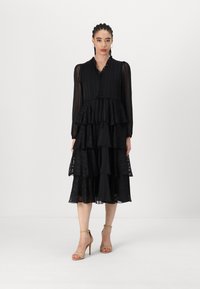 Black tiered dress with a buttoned front, ruffled neckline, and long sheer sleeves. Fabric appears light and textured. Worn with beige heels.