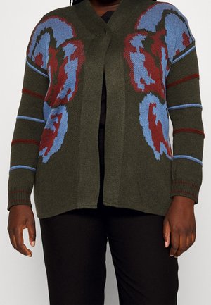 Person iført en åben, olivengrøn cardigan med blå og røde abstrakte blomsterprint og stribede ærmer, kombineret med sorte bukser.