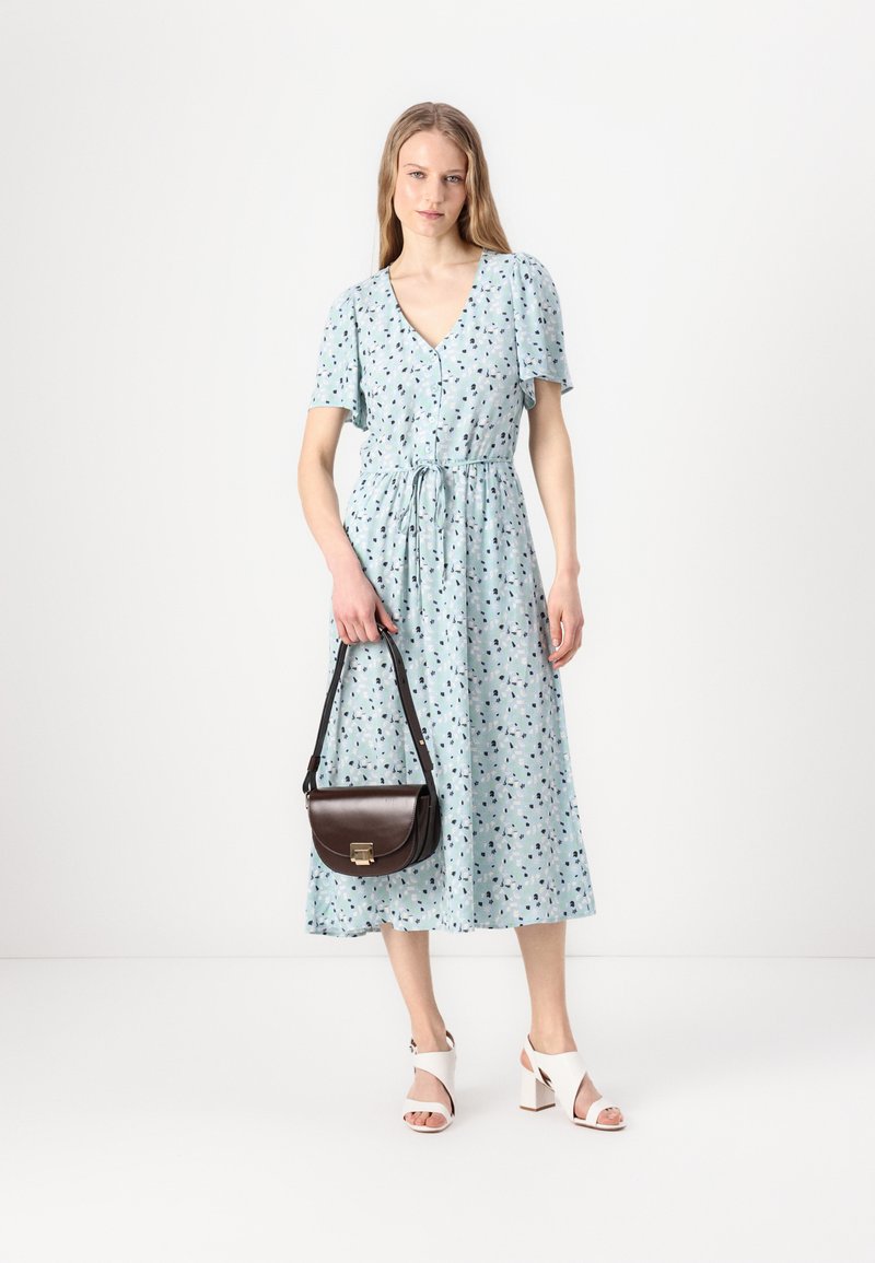Jasnoniebieska sukienka z krótkimi rękawami, wzór kwiatowy, dekolt w serek, ściągnięta w talii i do kostek. Brązowa torebka na ramię i białe sandały na obcasie.