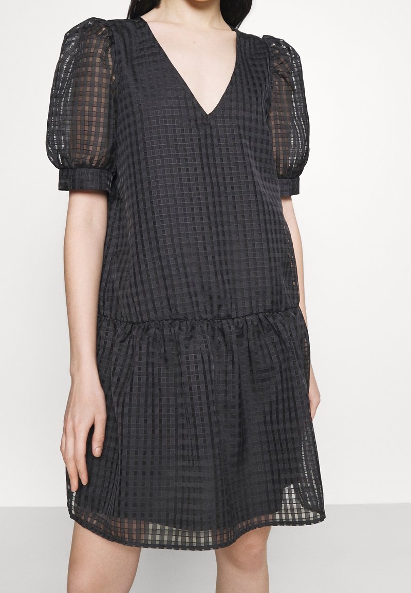 Femme portant une robe noire transparente à carreaux avec des manches courtes bouffantes et un décolleté en V profond, debout devant un fond uni.