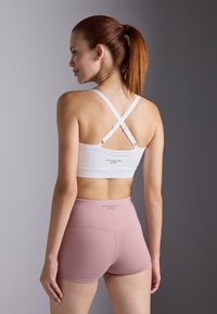 Sujetador deportivo blanco con tirantes cruzados en la espalda, combinado con pantalones cortos rosa de cintura alta. Ambas piezas presentan un diseño sin costuras y un logo de la marca.