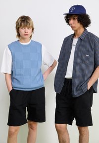 Hellblau strukturierte Weste über einem weißen Hemd, schwarze Shorts und eine blaue Mütze auf einer Figur; karierter Hemd auf der anderen Figur mit schwarzen Shorts.