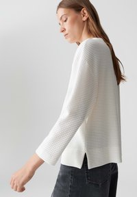 Giovane donna in un top bianco a maniche lunghe con texture e jeans neri, di profilo su uno sfondo grigio chiaro e uniforme.