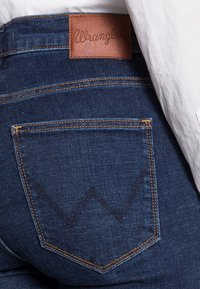 Mörkblå denimjeans med en läderetikett från Wrangler. Har bakfickor med kontrasterande guldstickning och ett distinkt "W"-mönster.