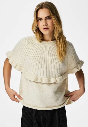 Donna con capelli biondi lunghi che sistema un maglione a collo alto color crema con dettaglio di mantella arricciata, indossando pantaloni neri a righe sottili.