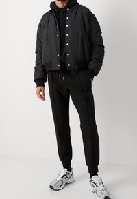 Svart kort jacka med tryckknappar, kombinerad med en svart hoodie och matchande joggers. Modellen bär silverfärgade sneakers med vita detaljer.