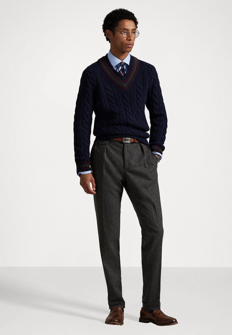 Maglione blu scuro a lavorazione cable-knit con scollo a V, pantaloni grigi, scarpe in pelle marrone e cravatta a scacchi. Testure sottili e accenti del maglione in contrasto.