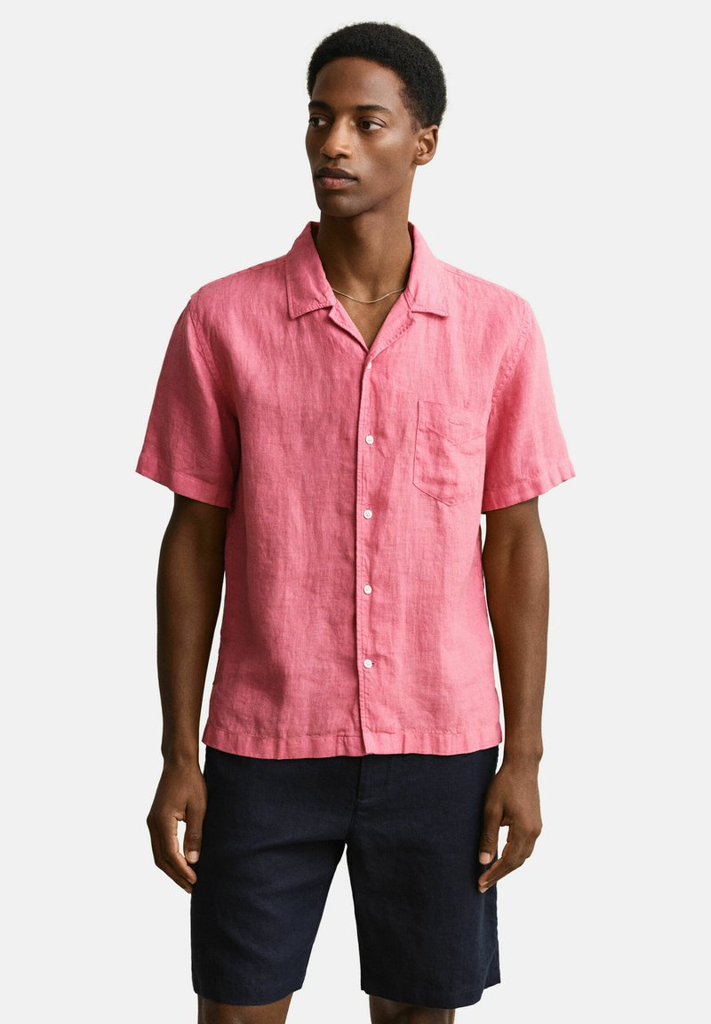 Homme portant une chemise boutonnée à manches courtes rose vif avec une poche poitrine et un short bleu marine foncé, debout devant un fond uni.