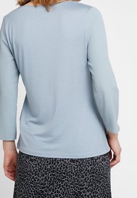 Světle modrý dlouhý rukáv s kulatým výstřihem, hladká textura a mírný lesk, v kombinaci s šedou sukní s černým leopardím potiskem.