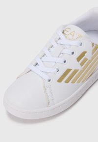 Hvit sneaker med gull geometriske striper, hvite lisser og branding på tungen, vist fra en skrå vinkel på en hvit bakgrunn.
