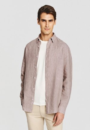 Młody mężczyzna w jasnobrązowej, rozpiętej koszuli na białym t-shircie i beżowych spodniach, stojący na jednolitym jasnym tle.