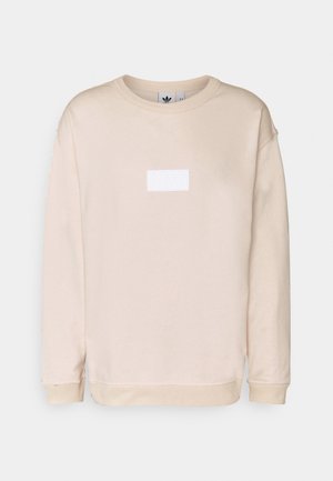 Homme portant une chemise beige à manches longues, un jean bleu clair retroussé, une ceinture noire, des chaussettes blanches et des baskets Adidas noires avec des bandes blanches.
