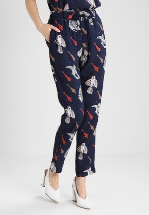 Pantalon bleu marine avec des motifs de poissons rouges, blancs et marron, porté avec des chaussures à talons blancs, une main dans la poche, debout sur un sol blanc.