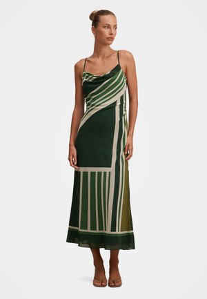 Kobieta w zielonej i beżowej sukience midi bez rękawów z geometrycznym wzorem oraz sandałach, stojąca lekko zwrócona w lewo na jednolitym białym tle.
