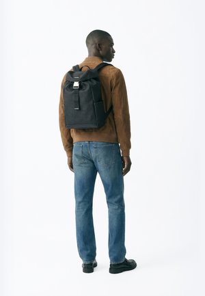 Mies, jolla on päällään ruskea takki, siniset farkut ja mustat kengät, kantaa mustaa reppua seisten sivuttain valkoista taustaa vasten.