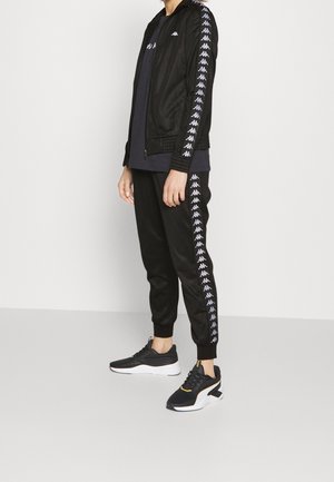 Personne portant un survêtement noir avec des logos blancs sur les manches et les jambes du pantalon, associé à des chaussures de sport noires et blanches, debout de profil.