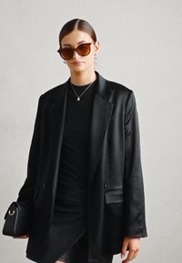 Blazer noir avec une coupe ajustée, texture lisse et deux boutons. Porté sur un haut noir et assorti à des lunettes de soleil sombres et un petit sac à main.
