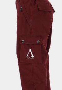 Bordeauxfarvede cargobukser med en frontlomme med klap og tryklås, blød tekstur og en hvid logo, der er tydeligt vist.