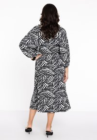 Schwarz-weiße Midi-Kleid mit Zebramuster, langen Ärmeln, gerafften Manschetten und ausgestelltem Schnitt. Getragen mit schwarzen High-Heels.