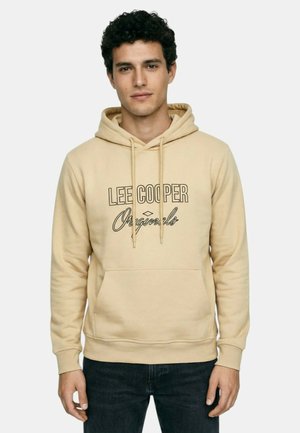 Jeune homme aux cheveux bouclés foncés portant un sweat à capuche beige Lee Cooper Originals et un jean foncé, debout devant un fond uni.