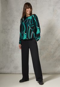 Blusa nera con un motivo astratto verde e bianco, abbinata a pantaloni neri a gamba larga e scarpe nere, il tutto su uno sfondo texturizzato.