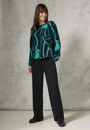 Blusa nera con un motivo astratto verde e bianco, abbinata a pantaloni neri a gamba larga e scarpe nere, il tutto su uno sfondo texturizzato.