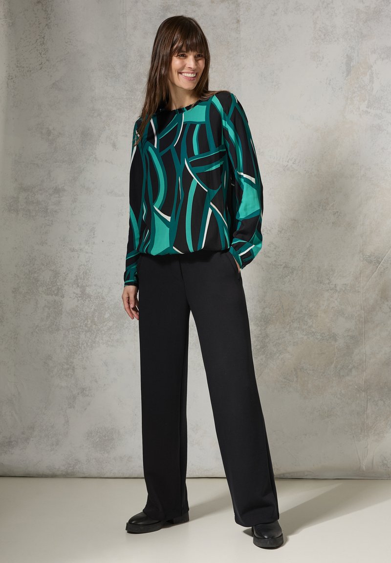 Blusa nera con un motivo astratto verde e bianco, abbinata a pantaloni neri a gamba larga e scarpe nere, il tutto su uno sfondo texturizzato.
