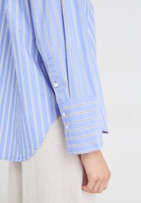 Camicia a maniche lunghe azzurro chiaro con strisce bianche, caratterizzata da polsini con bottoni e una sottile texture, abbinata a un tessuto di colore chiaro sotto.